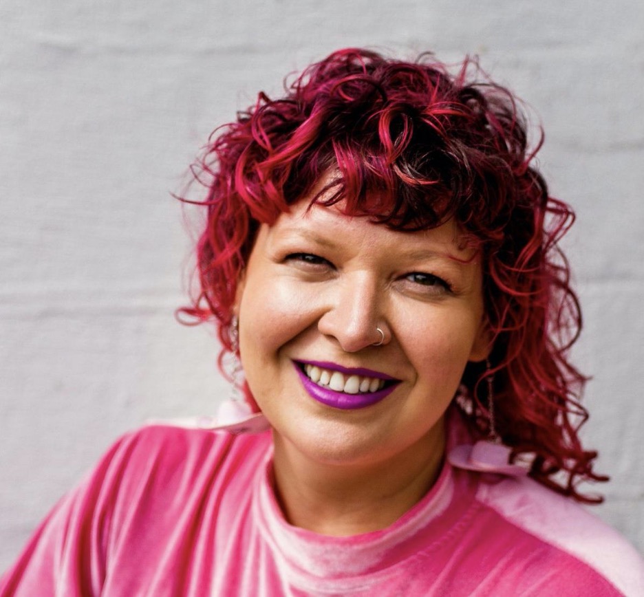 Dr. Jessie Waggoner, a person with pink curly hair and wearing a pink velvet shirt smiling for the camera. Behind them is a white brick wall.